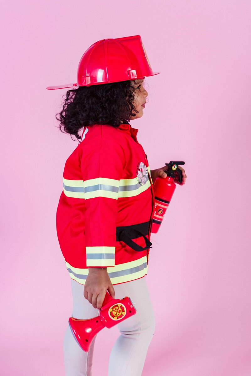 A young child dressed in a bright red firefighter costume, complete with a red helmet, is holding a toy fire extinguisher and a red megaphone against a pink background.