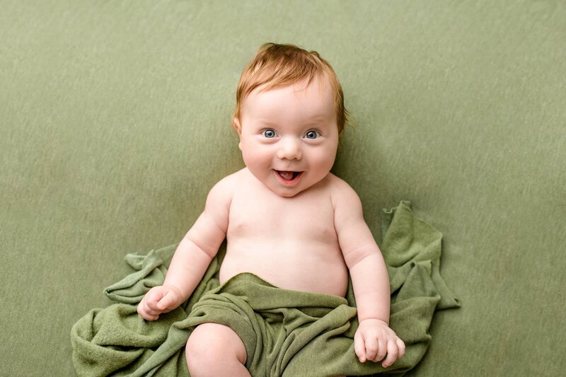 four month old baby boy lays on green background in his milestone portrait session with cloth around him smiling with his bright blue eyes and red hair standing out