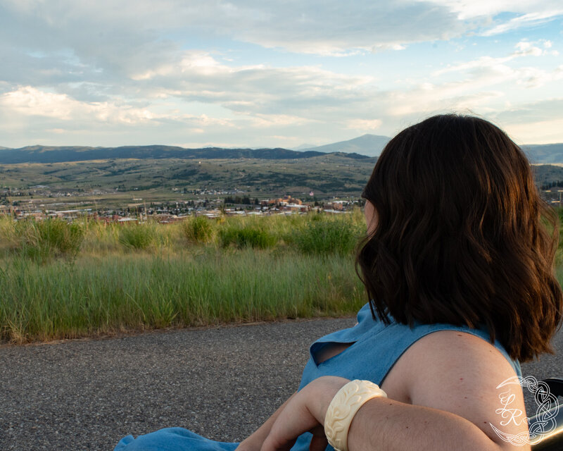 photo of Ashlyn standing on a sidewalk in a blue denim dress, tossing her hair over her shoulder and smiling. alfalfa, grass and headframes int he background