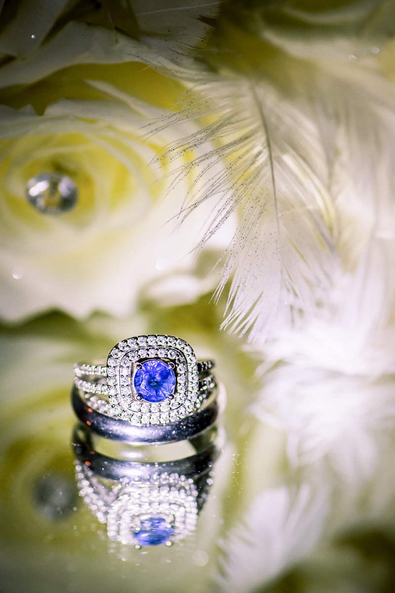 Sapphire and diamond wedding rings sit on mirror with feathers and yellow rose in background