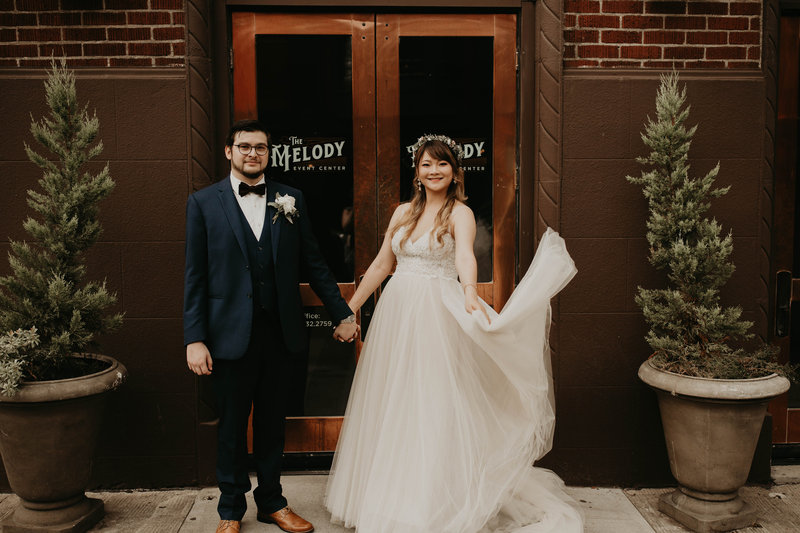Bride swirls wedding gown skirt while holding hands and smiling with her groom in front of The Melody in Portland, Oregon