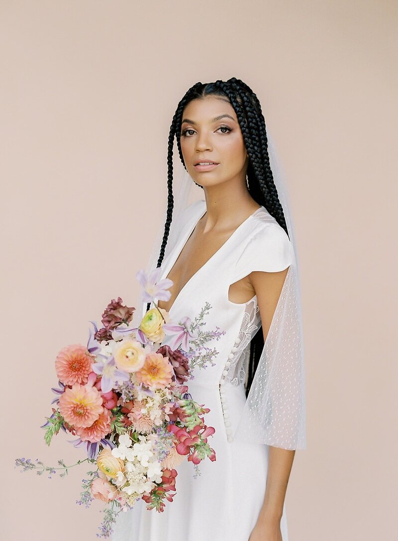 Black bride with long braids in a white dress and veil holding a colorful bouquet with dahlias and ranunculus