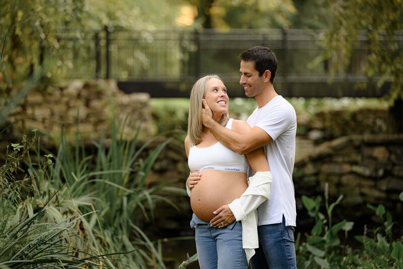 a soon to be father touches his wifes face as they stand in a garden during materntiy session