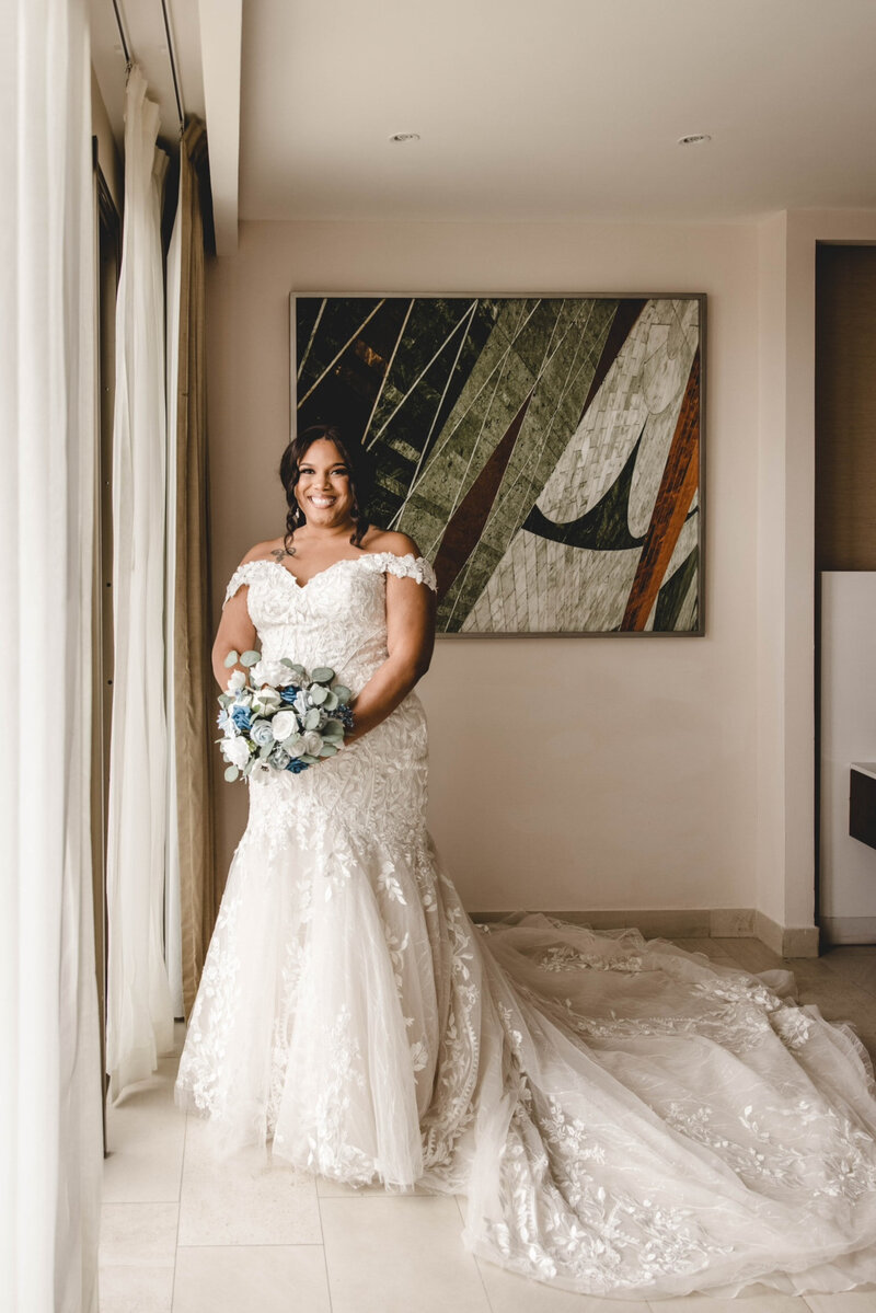 Bride smiles in her wedding dress next to a window.
