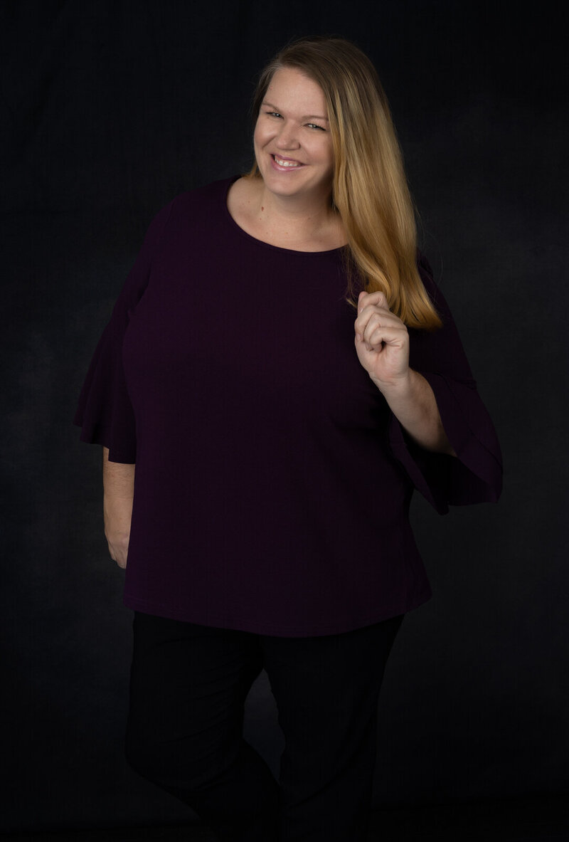 photographer-headshot-wearing-purple-shirt-in-studio-with-hand-on-hip-holding-blue-chair-in-arlington-tx