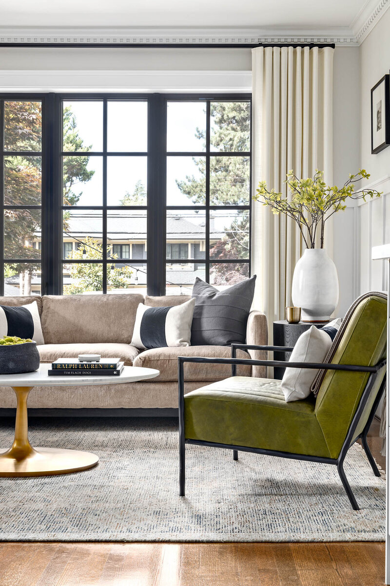 Living room design, there is a large window with black beans, a beige sofa, a grey woven rug, and a sued green seating chair with a black frame. The living room feels airy and mid-century modern in style