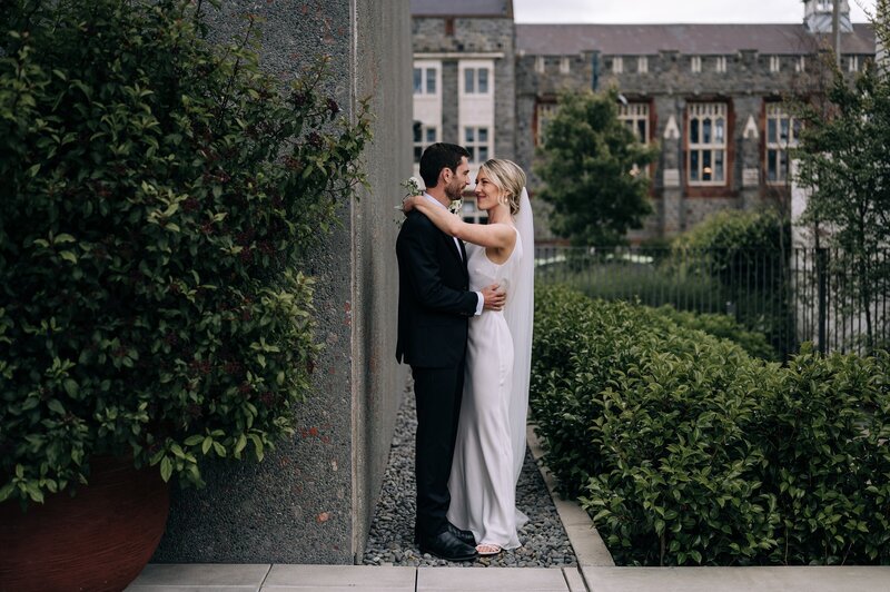a bride and groom with their arms around each other between a concrete building and low bushes with the canterbury museum in the background nz