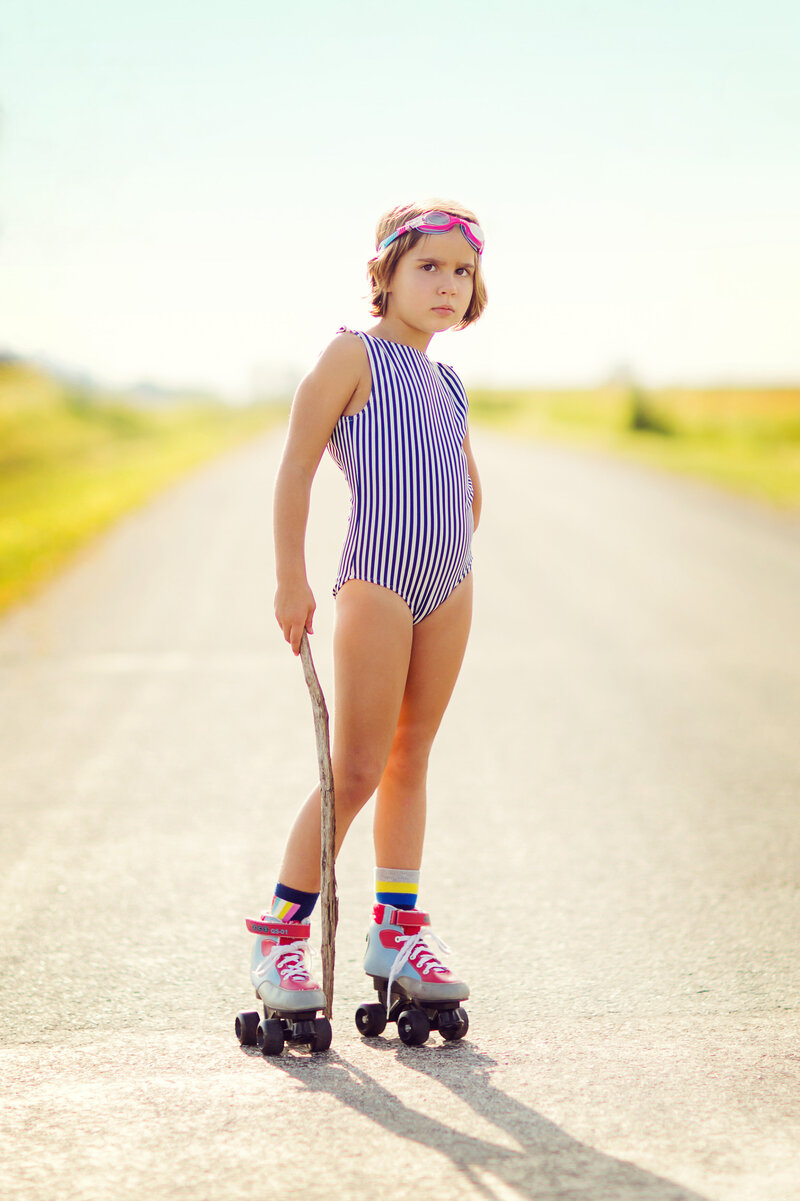 Young girl in a blue and white striped one piece bathing suite, holding  a long stick, with swim goggles, striped socks, and roller skates, on a country road.