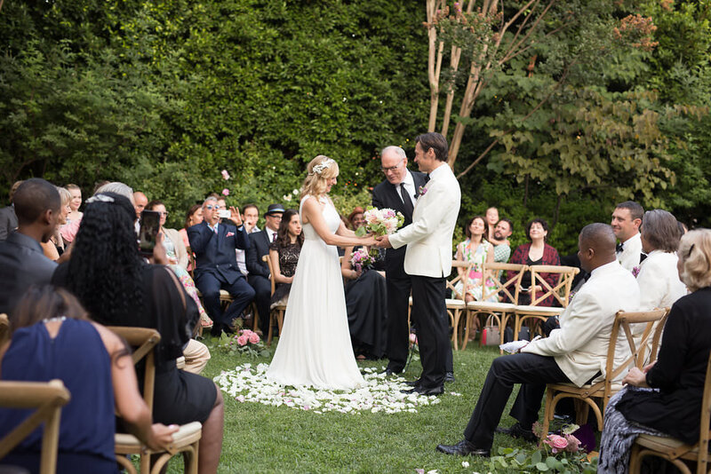 Bride and groom take their vows atop a cirle of white rose petals surrounded by guests