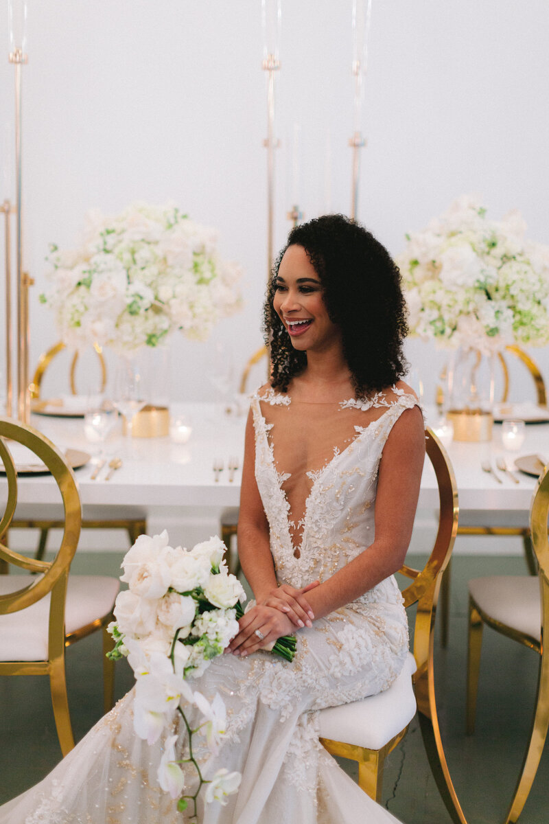 Bride smiles while holding white orchid floral bouquet, sitting at wedding reception table