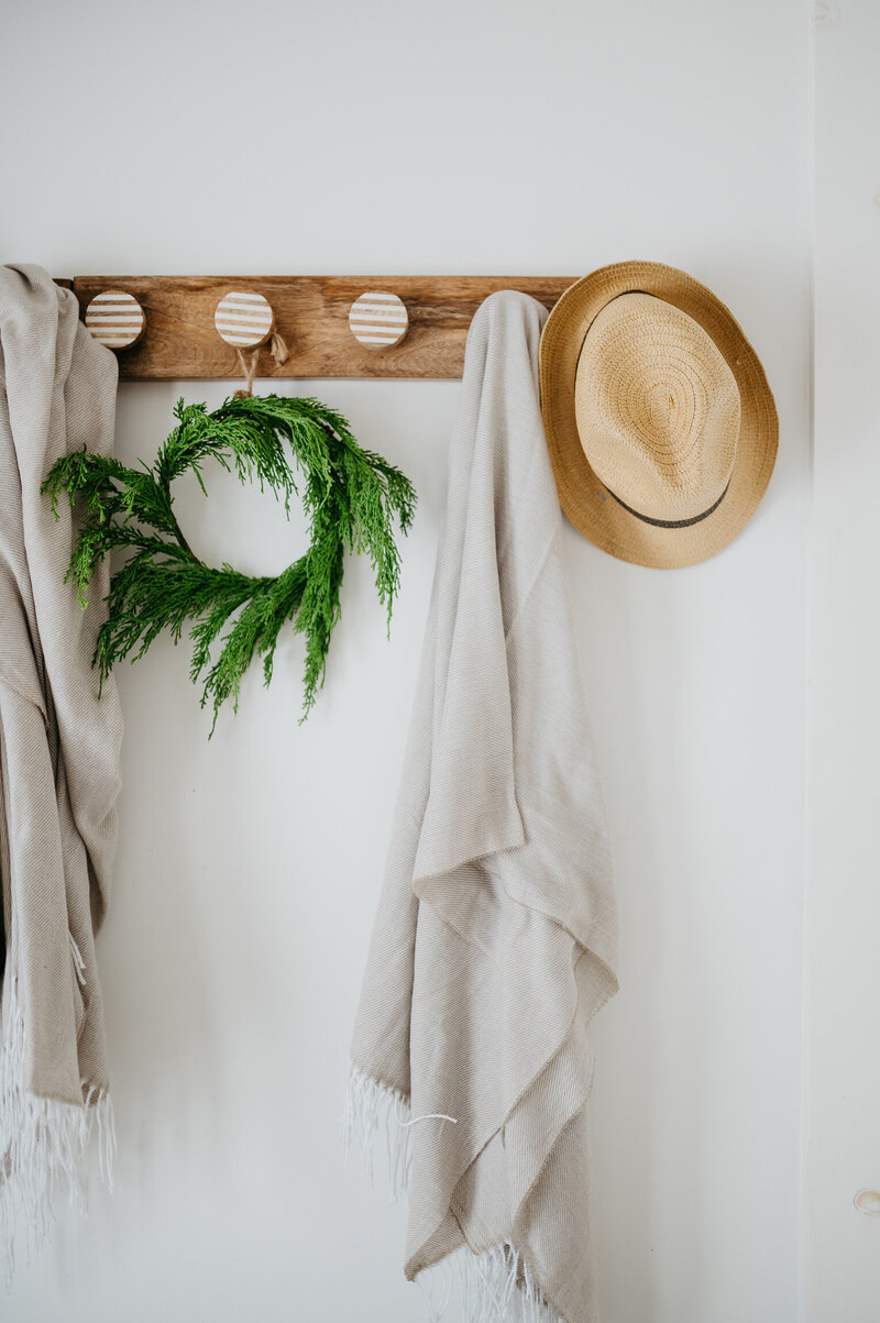 Minimalist home styling with natural greenery wreath and woven hat hanging on wooden peg rail with white linens.