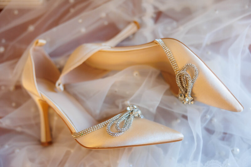 Wedding Day jeweled shoes photographed with white lace background.  Details of the Brides shoes and how they look before she wears them for her ceremony.  Photography by Angela Foushee, a Virginia Wedding Photographer  Diamond bow accents on shoes.