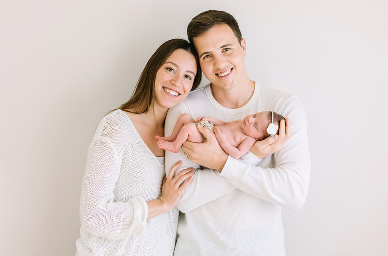 A new family dressed in white smiles at the camera holding their newborn baby daughter