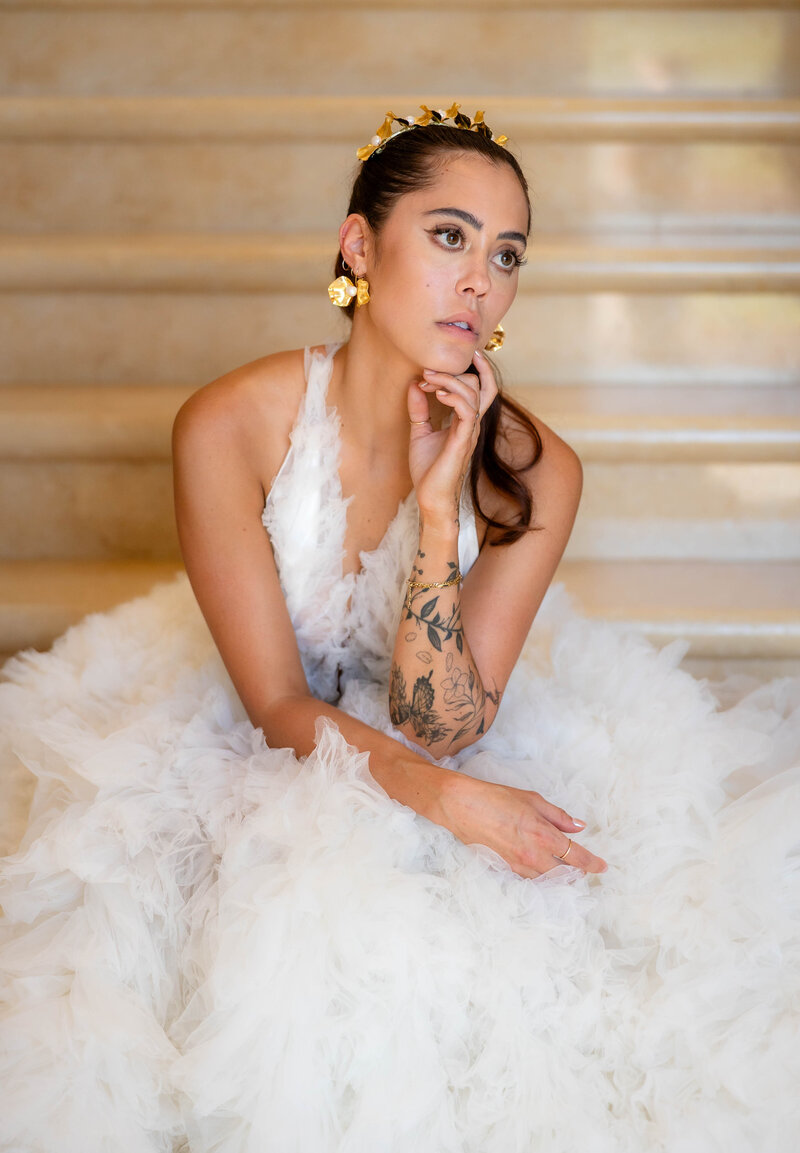 Bride in a Millia London type gown with plunging neckline and soft tulle ruffles skirt.  Bride is sitting on the  bottom stair of the marble staircase at the wedding venue