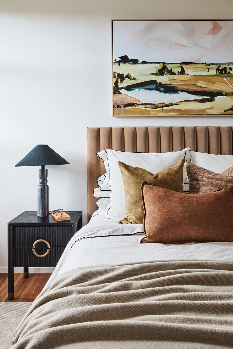 Contemporary bedroom with tan upholstered bedhead, layered cushions, and black bedside table with modern lamp.