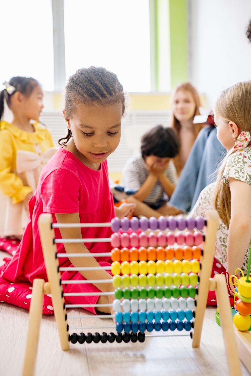 A group of children are playing together, with one child in a bright pink dress focusing on a colorful wooden abacus, while other children and a teacher interact in the background.