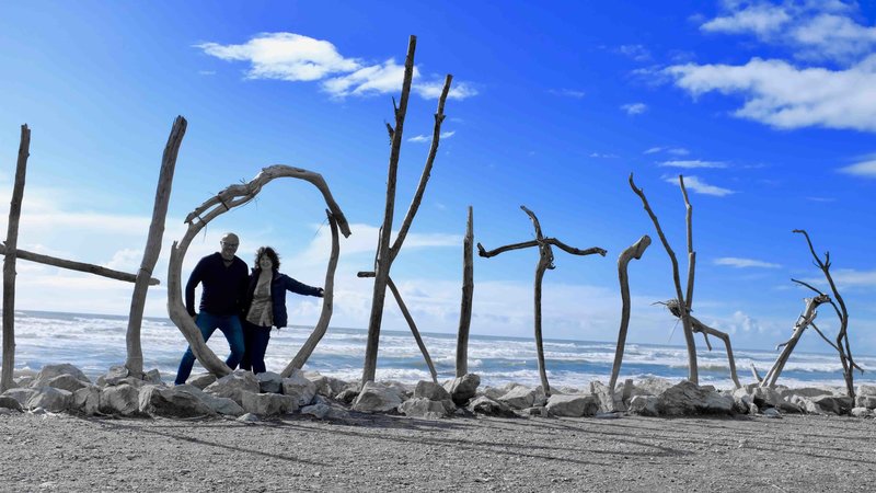 Lynda and Warren standing behind the Holitika sign made from driftwood on the beach in Hokitika, South Island, New Zealand