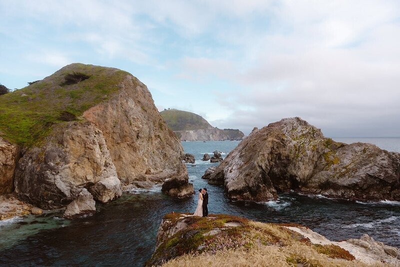 A couple stands close together on a rocky cliffside above the ocean, surrounded by dramatic coastal rock formations and waves, with green hills and cloudy skies in the background in Big Sur, California.