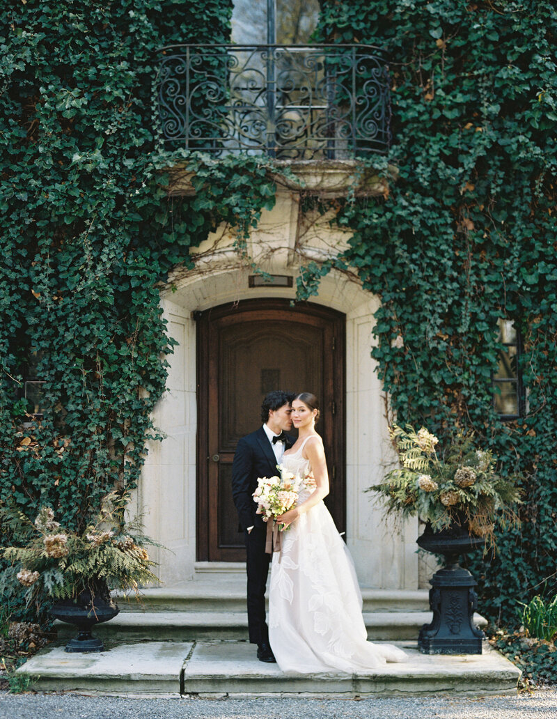 Bride and groom in front of brown door with black tux and Berta wedding gown.