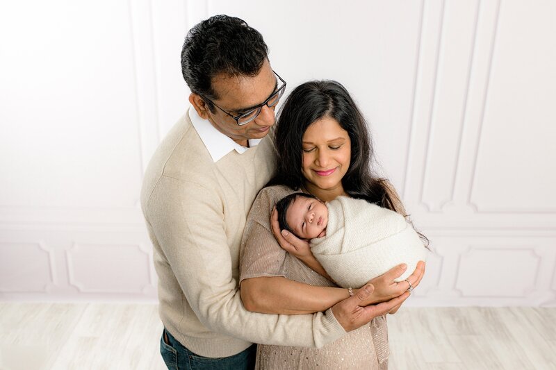 mother and father are both dressed in neutral tones with mother holding a newborn son wrapped and cream looking down towards him smiling