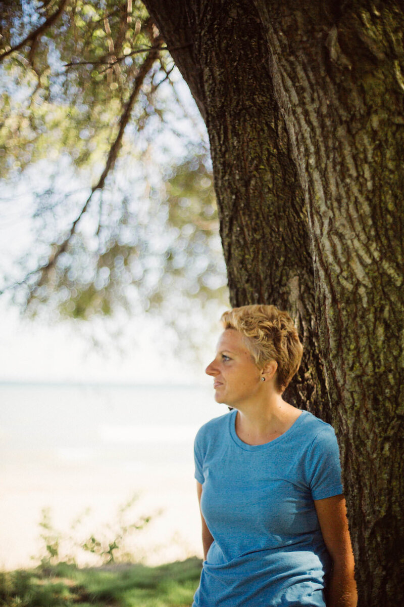 Writer, Sarah Freymuth, leans against a tree and looks off into the distance in a blue t-shirt