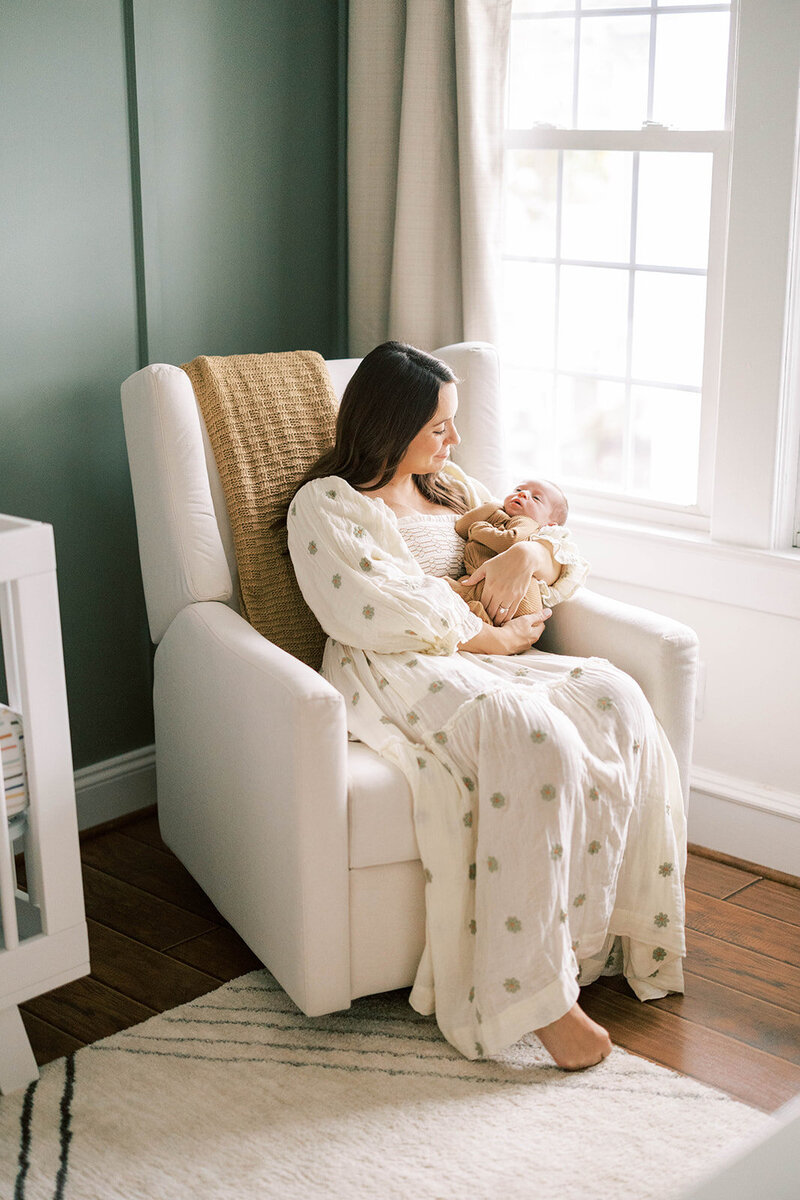 Mom sits in white nursery chair to cradle sleepy newborn baby at their Mechanicsburg home.