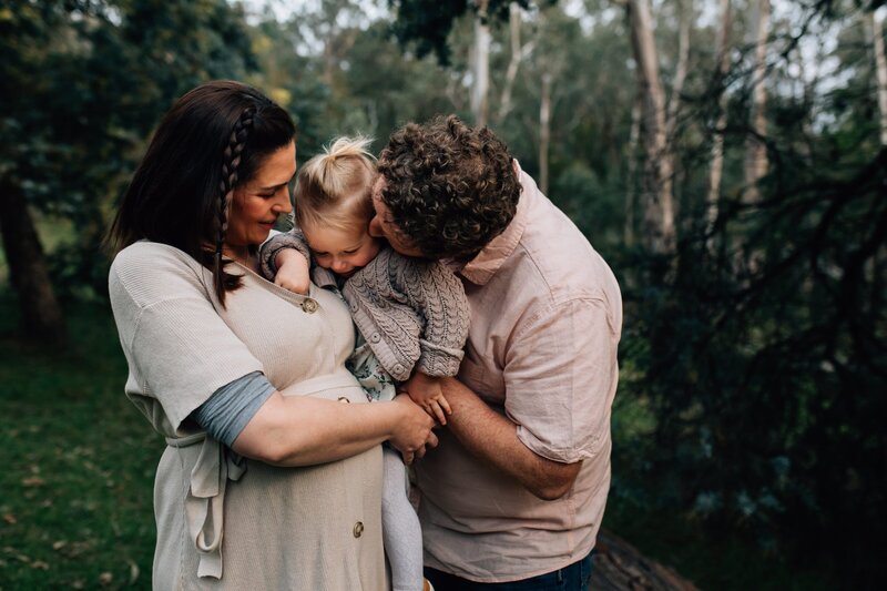 Mum, Dad and child cuddling together outside in bushland  during maternity photography session.