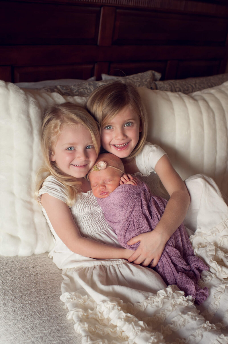 Two older girls snuggling their newborn baby sister on the bed for their lifestyle in home session