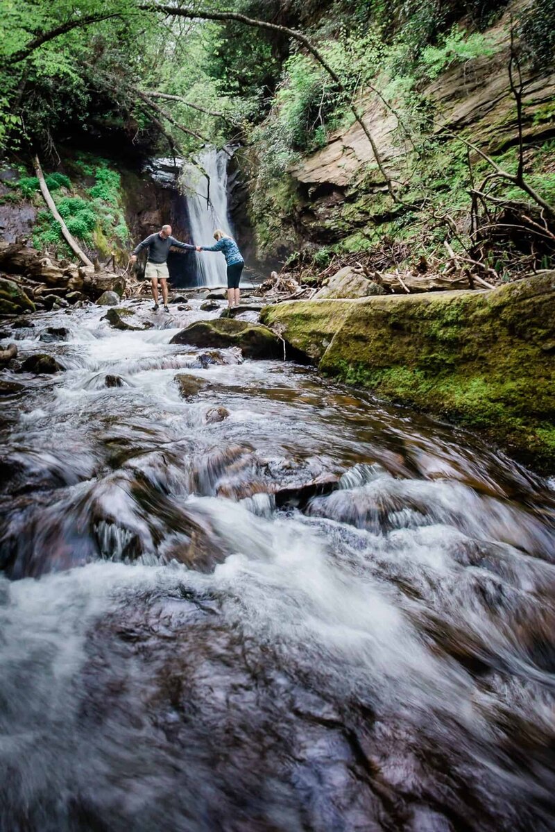 On day one of their luxury destination elopement with their photographer, a couple is out exploring where their ceremony will be the next day. It is a lush green hollow in the South, filled with a stream with little rapids smoothly flowing with water and the couple carefully crossing on large rocks further up stream with water fall behind  them. 