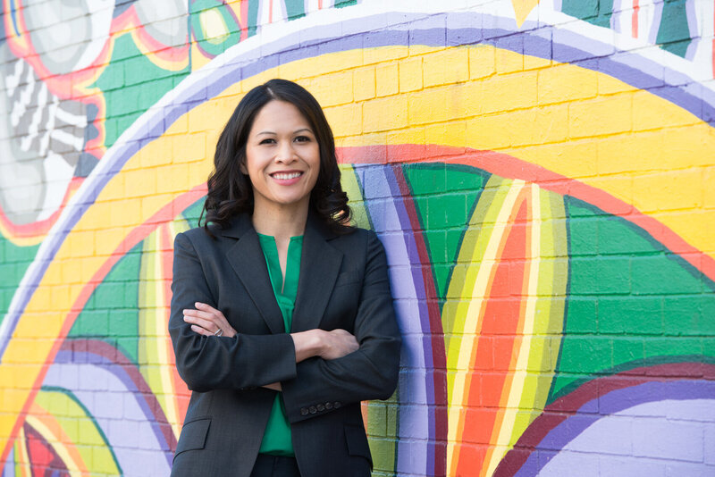 Dr. Jen, a Filipino woman with shoulder-length dark hair, wears a green blouse and dark blazer over top. She is leaning against a colorful mural painted on a brick wall behind her, smiling at the camera.