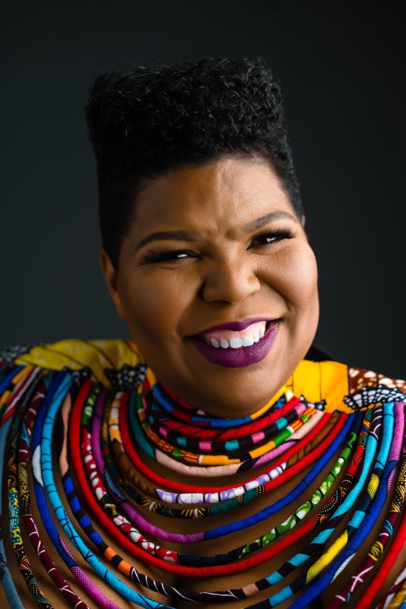 A vibrant portrait of a woman smiling brightly, adorned in a colorful, layered neckpiece made from various African fabrics