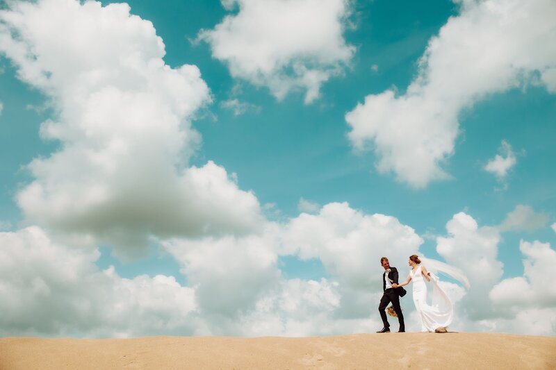 A bride and groom walking along the sand with a bright blue sky full of clouds behind them. Captured by Infinite Productions