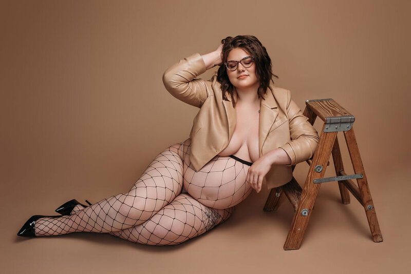A pregnant woman in a light leather jacket, fishnets, and heels sits on the floor leaning against a wooden ladder in front of a neutral tan solid backdrop, captured by a photographer in Bemidji, Minnesota