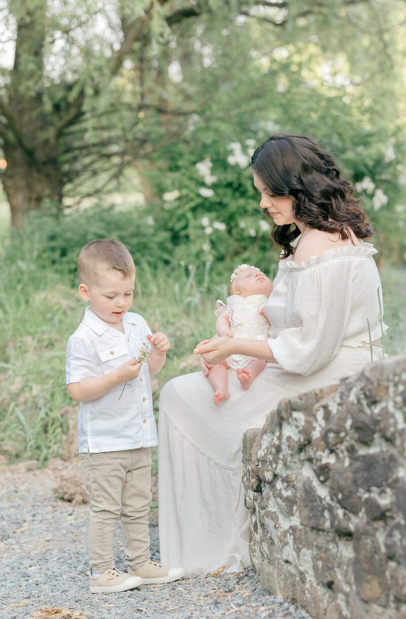 A mom sitting on the side of a small stone bridge with her newborn baby and her toddler son with white flowers in the background by NJ Family Photographer