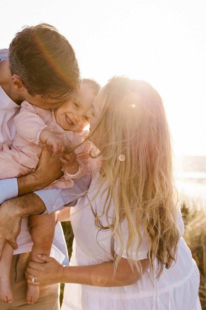 A mother and father holding their giggling baby during their sunset beach photoshoot with aimee kelly in taranaki