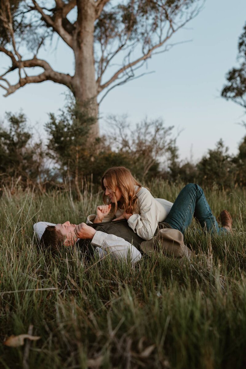 Romantic couples photography capturing a pair lying in a grassy field, smiling and embracing in photography session.