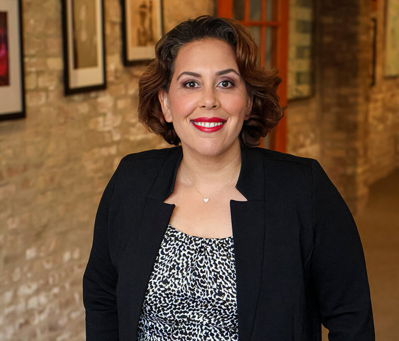 Smiling person with short brown hair, wearing a black blazer and patterned top, stands against a brick wall with framed artwork. Warm, welcoming tone.