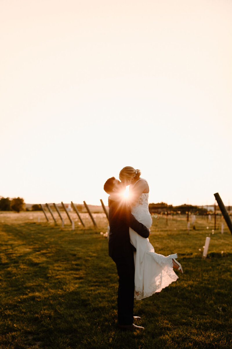 Gorgeous wedding photoshoot by Morgan Ashley Lynn Photography in Lake Country, Wisconsin out in a field at sunset with groom picking up the bride and kissing her, the sun is perfectly placed right in between them