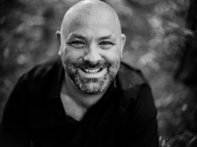 Portrait of a smiling bald man with a beard, wearing a black shirt against a blurred natural background