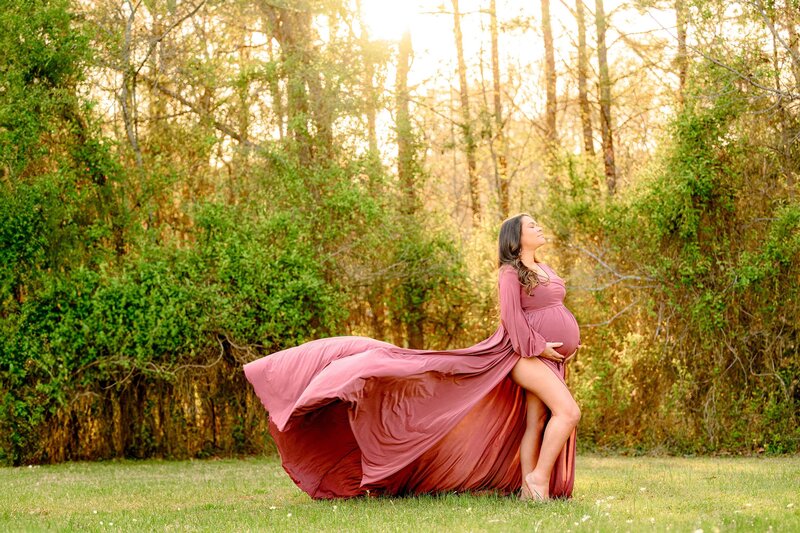 Charlotte Area mom is posed with one light out and a rose colored maternity dress with the train flowing in the Wind and her face pointed upward towards the Sun