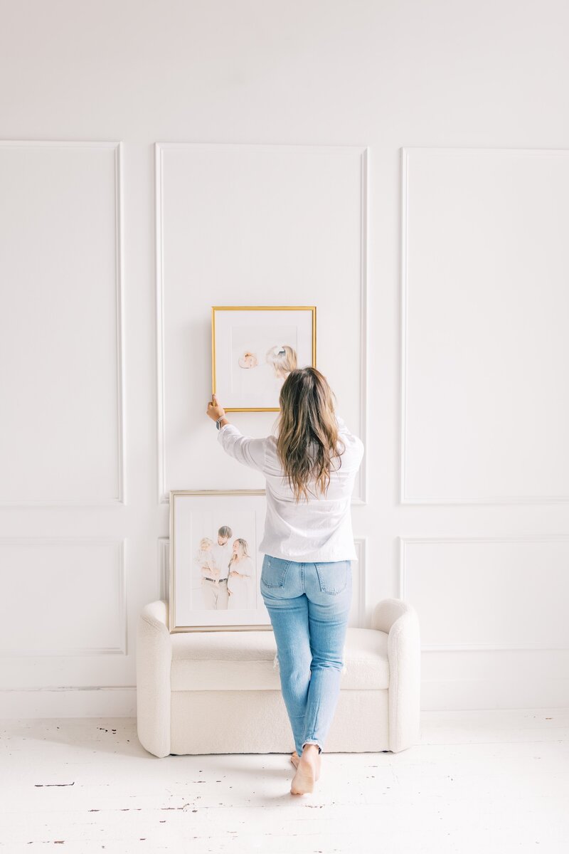 photographer standing in a white room with framed pictures.