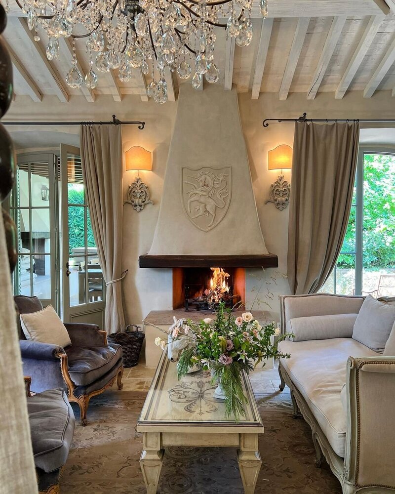 The Tuscan chic living room of a guest suite at Borgo Santo Pietro with a tromp l'oeil crest on the stone fireplace, antique wooden furniture and velvet armchairs