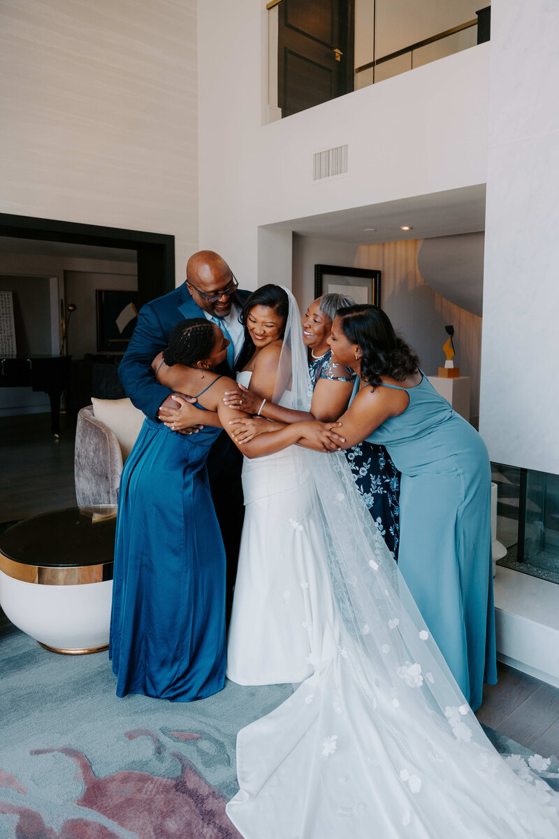 A group of five women and one man, likely family members, are embracing a bride in her wedding dress and veil. They are all smiling and appear happy. The scene takes place indoors, possibly in a hotel suite or a spacious home, with modern decor.