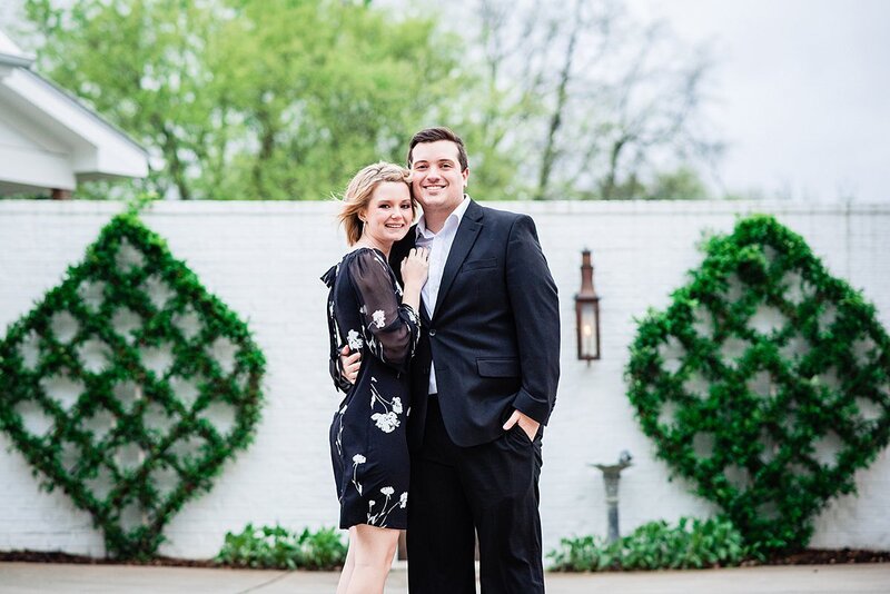 The bride and groom embrace in front of a white fence with green lattice topiaries in the background. The groom is wearing a charcoal gray suit with a white shirt and the bride is wearing a short dress with long sleeves and a high neckline. They are both smiling at the camera.