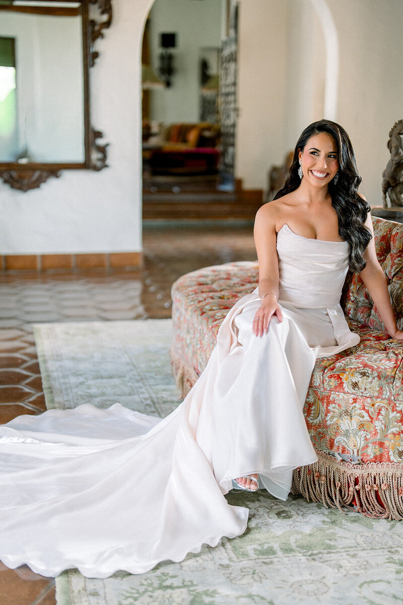 Bride smiles while seated on vintage ottoman in elegant bridal suite before luxury wedding in Southern California