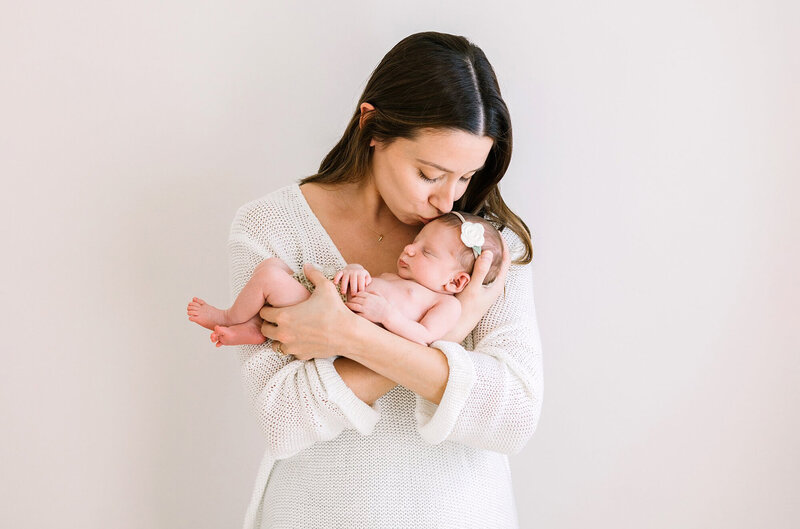 A new mom wearinga white dress kisses her newborn baby girl on the head