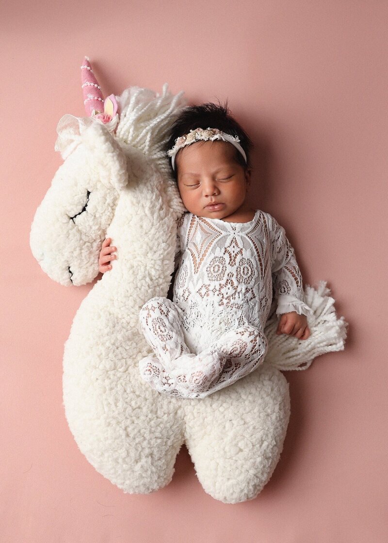 Sleeping newborn posing on pink blanket cuddling a white unicorn prop