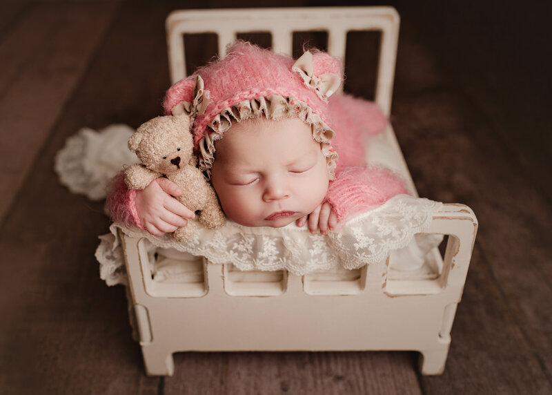 Newborn baby girl in bed pose holding a bear with a bear hat