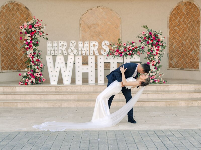bride and bridal kissing in front of marquee letters covered with bright spring flowers