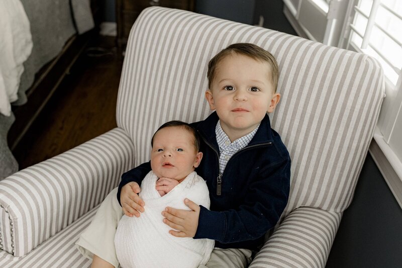 older son holds his baby brother wrapped in white while he is dressed in a Navy sweater and pants sitting in a chair